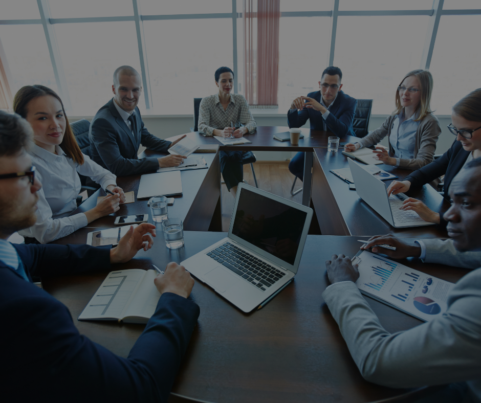 Un gruppo eterogeneo di professionisti in abiti formali è seduto attorno a un tavolo da conferenza, collaborando con computer portatili e grafici.