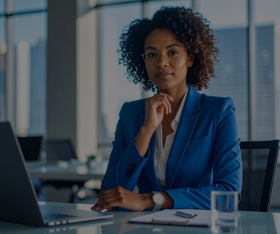 Professionista con capelli scuri e ricci, indossa un blazer blu ed è seduta a una scrivania in ufficio con un computer portatile e dei documenti.