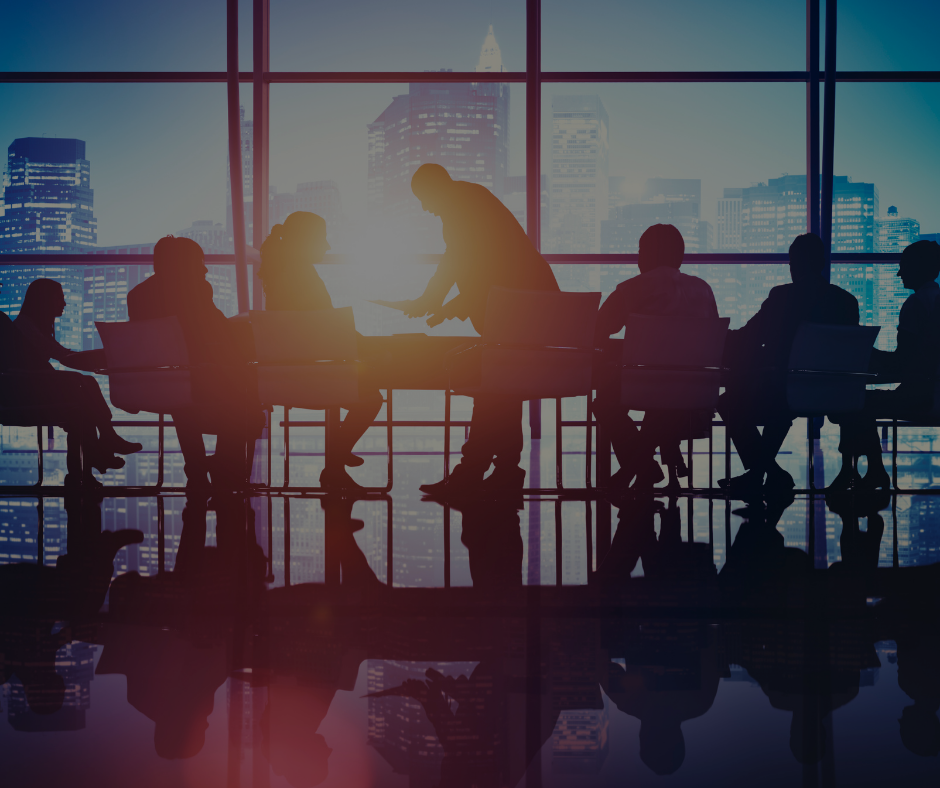 Le sagome di alcuni professionisti del mondo degli affari sono sedute attorno a un tavolo da conferenza di fronte a una grande finestra che si affaccia su una città al crepuscolo.