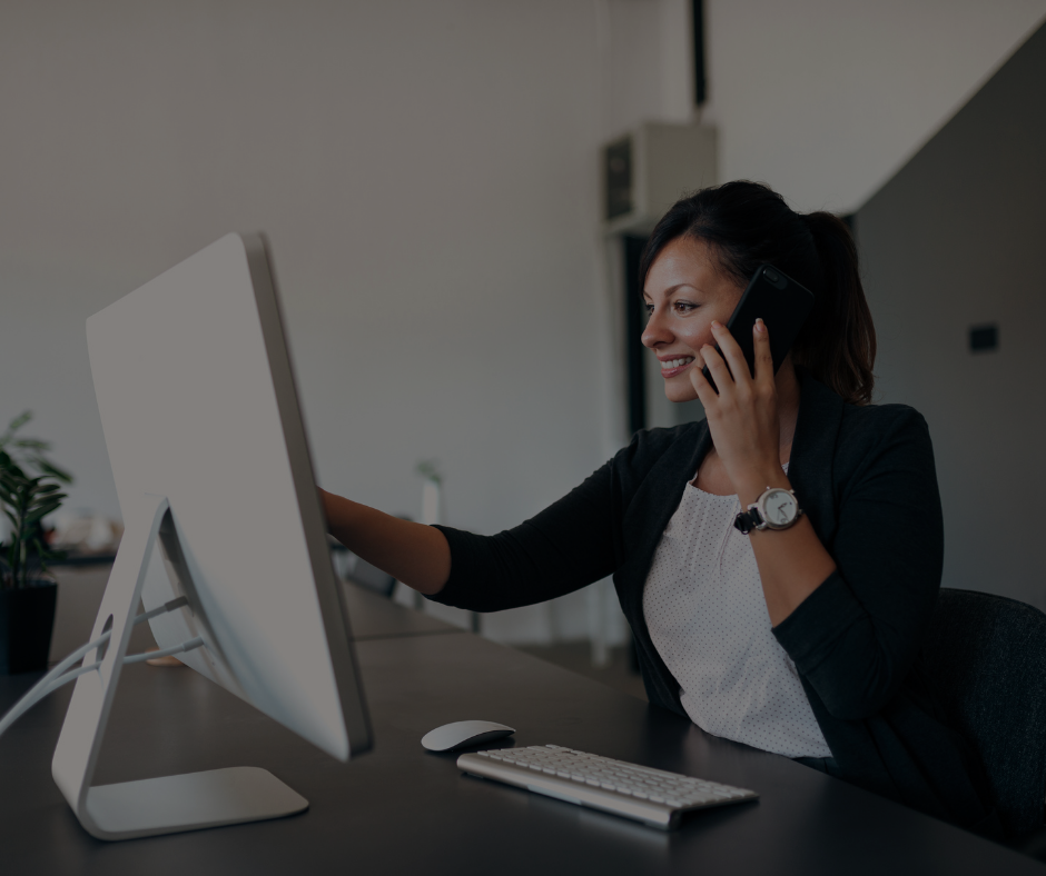 Un professionista al lavoro alla scrivania sorride mentre parla al telefono e indica lo schermo di un computer fisso.