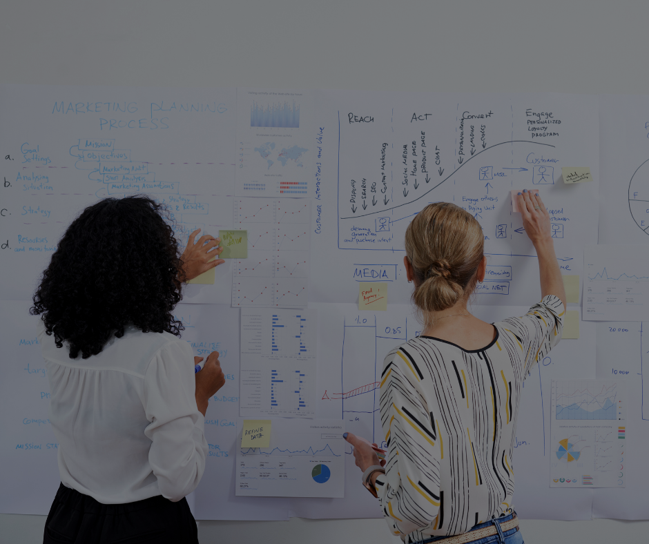Due persone in piedi davanti a una lavagna ricoperta di grafici e appunti, intente a organizzare e analizzare dati in modo collaborativo.