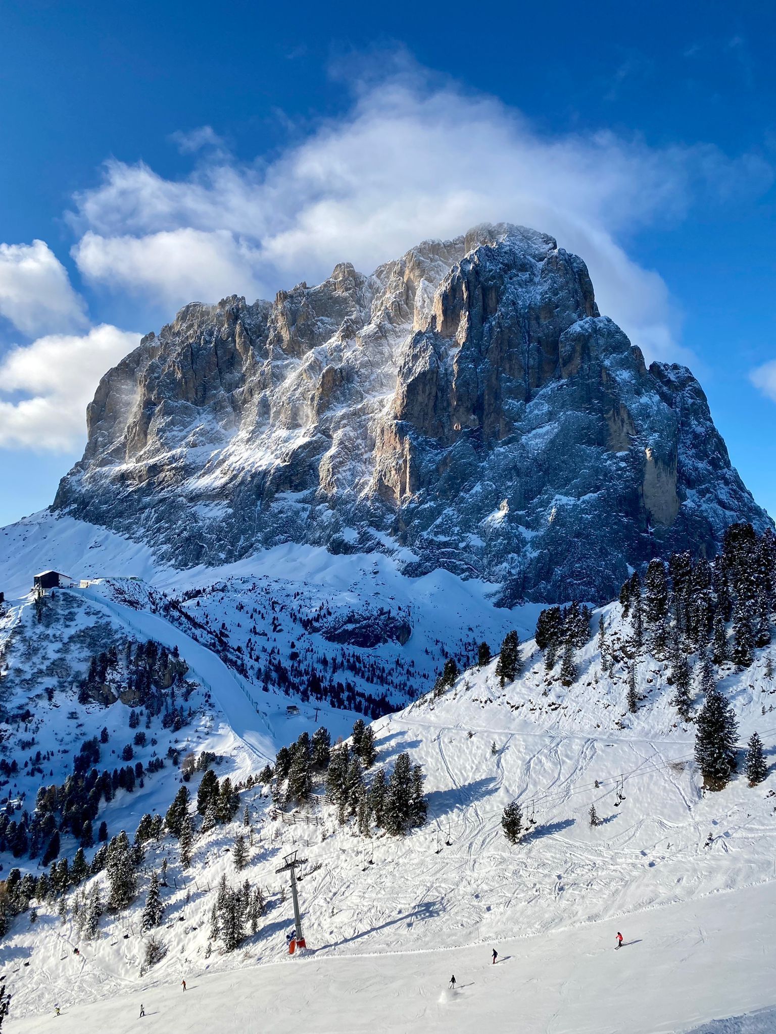Un'imponente vetta innevata si erge sopra una luminosa pista da sci soleggiata, punteggiata da pini e dotata di un impianto di risalita.
