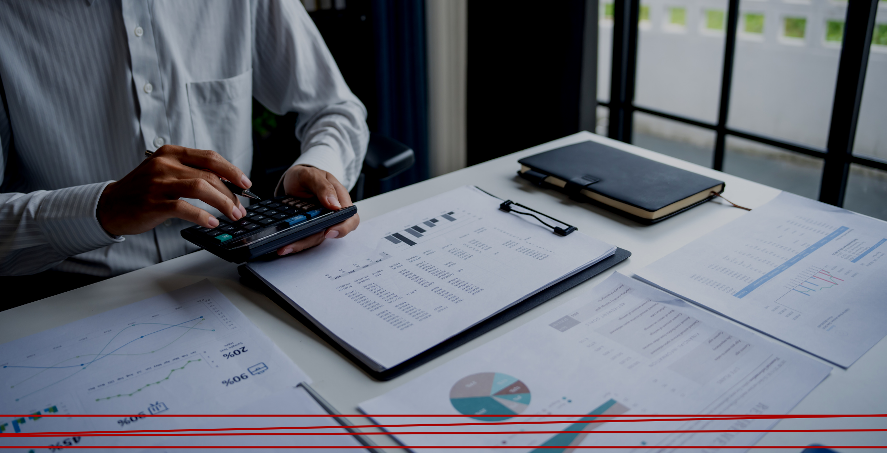 Una persona che usa una calcolatrice a una scrivania, con documenti finanziari, grafici e un quaderno.