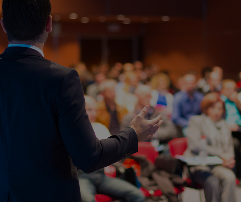 Una persona in abito scuro si rivolge a un pubblico seduto in un'aula magna.