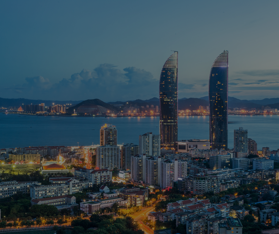 Al crepuscolo, due grattacieli gemelli dalle linee curve si ergono sopra il fitto skyline di una città costiera, affacciati su un porto con le colline sullo sfondo.