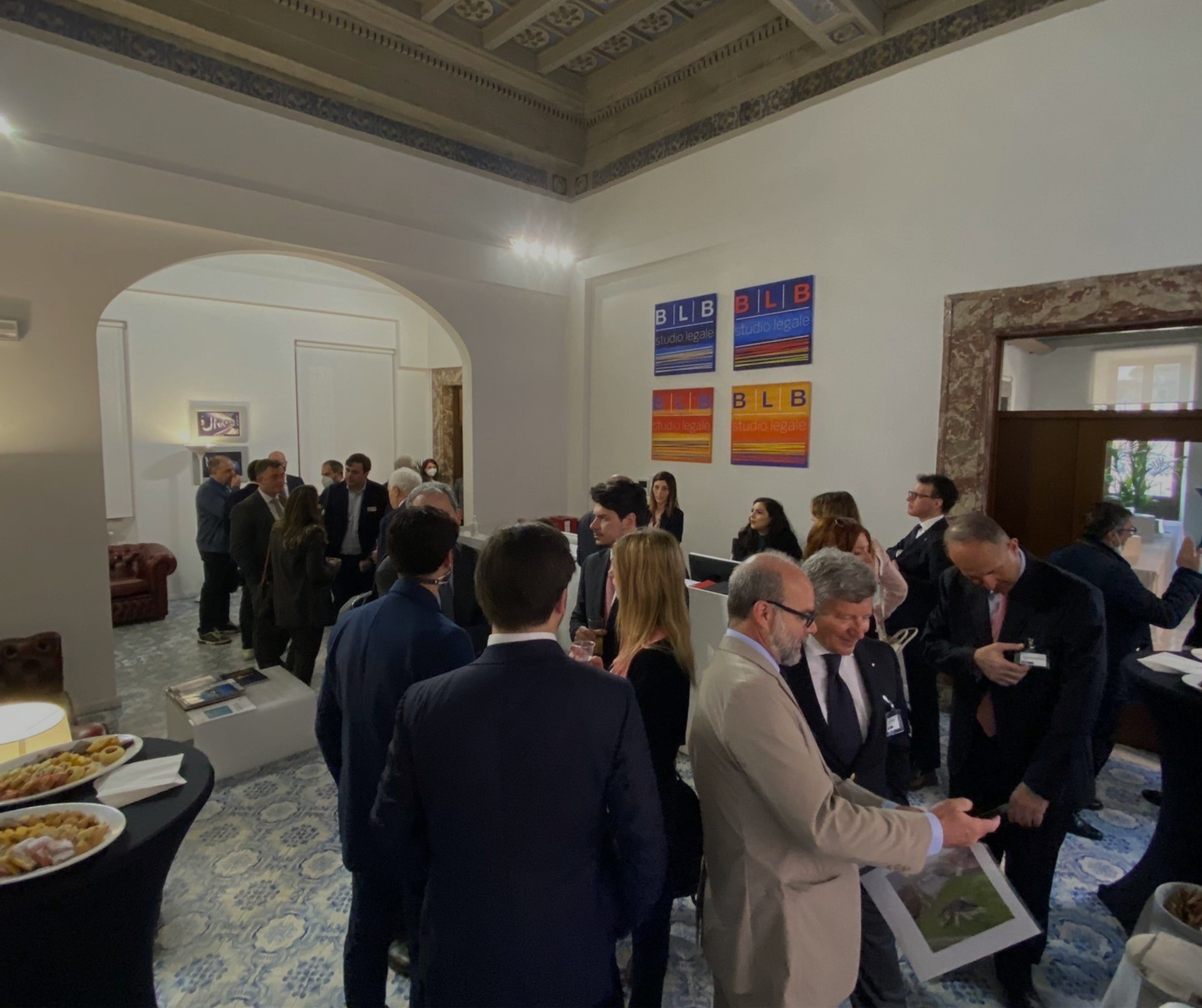 I partecipanti socializzano durante un evento di networking al coperto, tenuto in un'ampia sala con soffitti alti e opere d'arte incorniciate alle pareti.