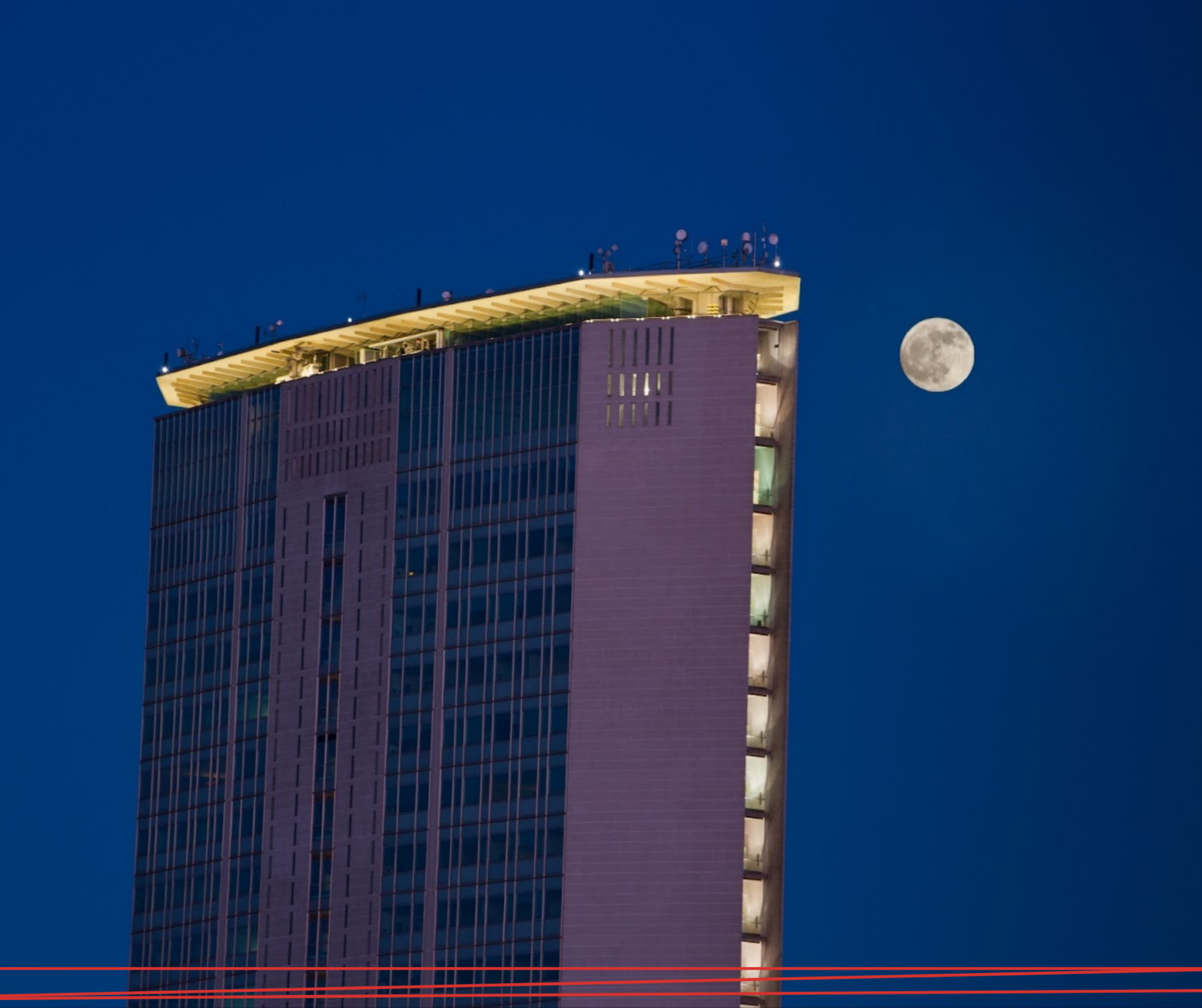Un grattacielo moderno con il bordo del tetto illuminato, stagliato contro un cielo serale blu scuro con la luna piena nelle vicinanze.