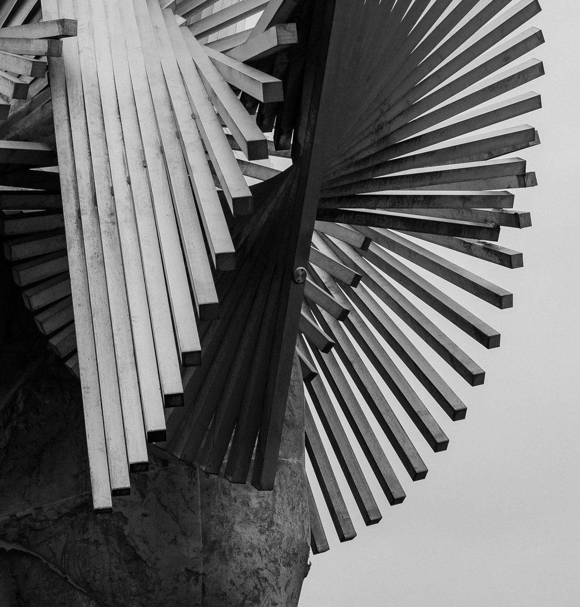 Una fotografia in bianco e nero, scattata dal basso, di una scultura realizzata con sottili listelli di legno sovrapposti e disposti secondo uno schema radiale a spirale.