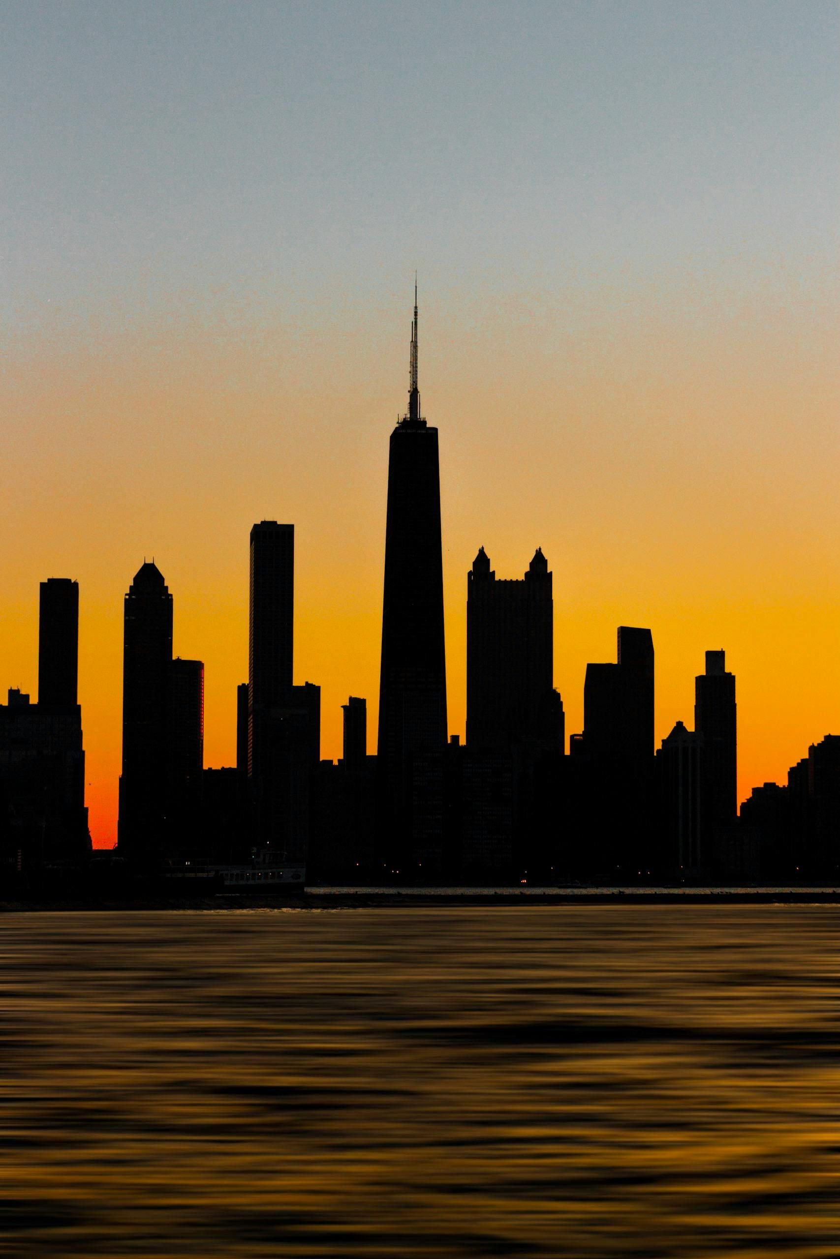 La sagoma dello skyline di Chicago si staglia contro un tramonto arancione, riflesso nell'acqua sottostante.