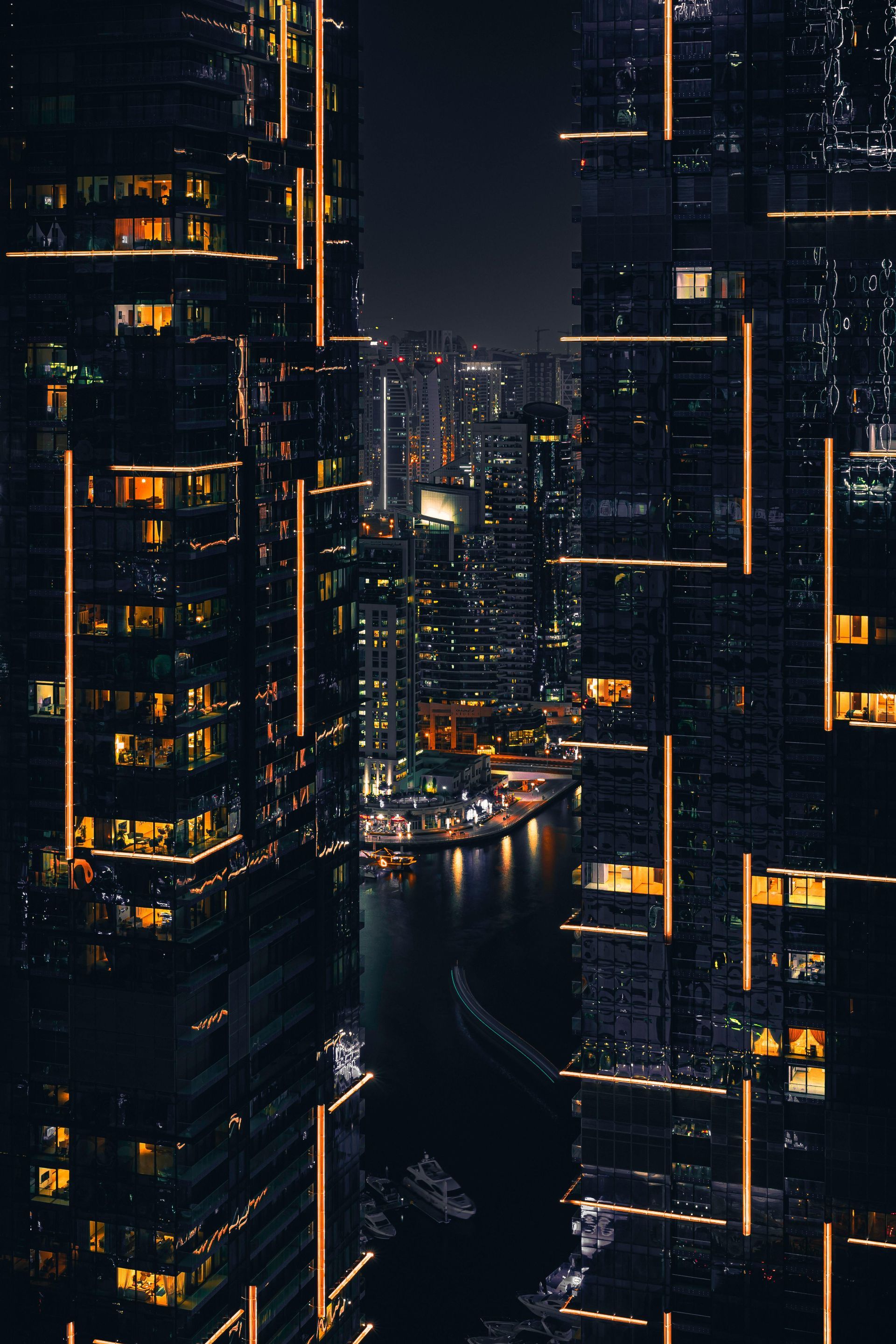 Di notte, alti e moderni grattacieli incorniciano uno stretto canale fluviale illuminato, riflettendo le luci della città in un paesaggio urbano oscuro.
