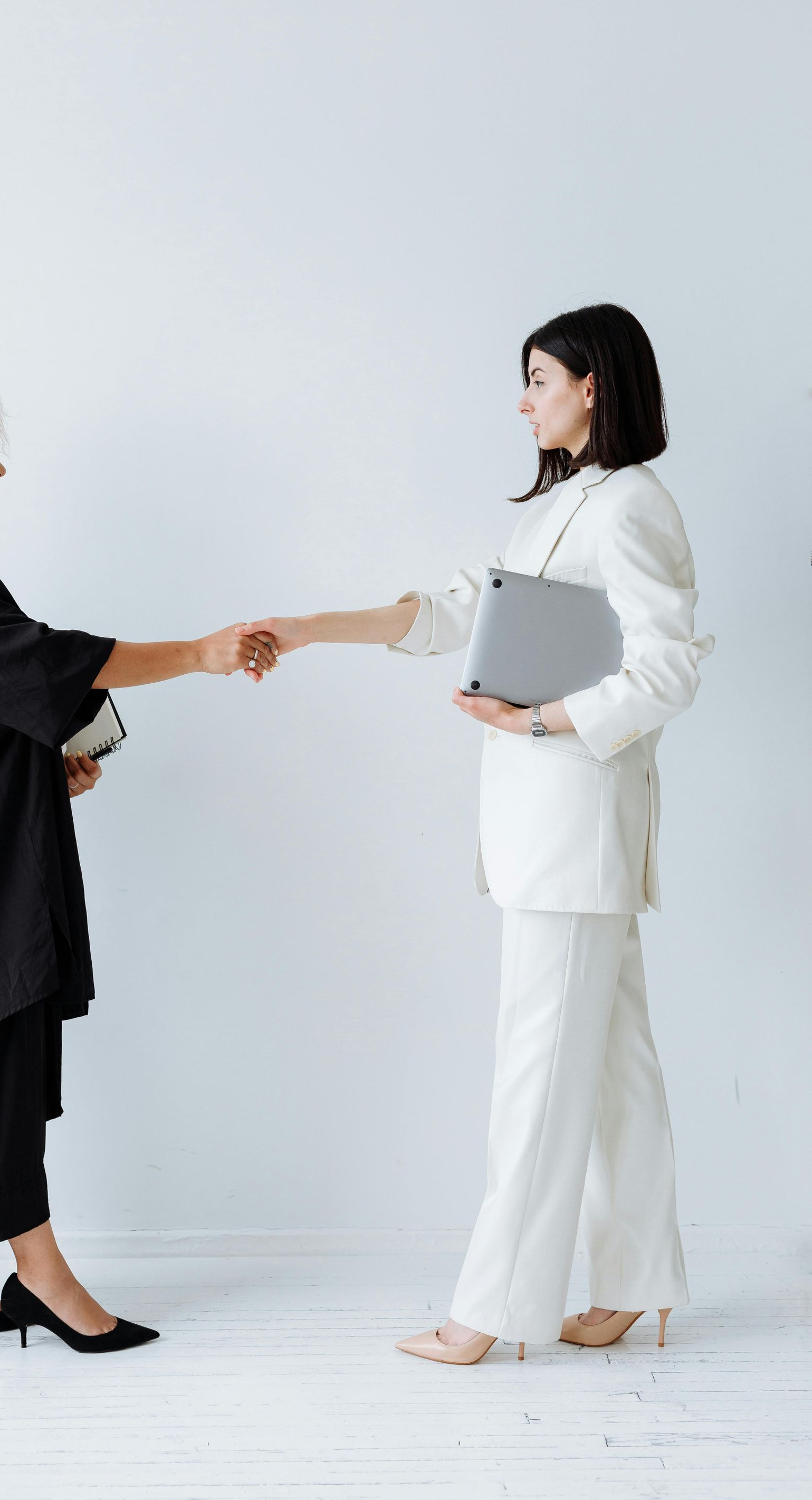 Due persone in abiti professionali, una in nero e una in bianco, si stringono la mano su uno sfondo bianco uniforme.