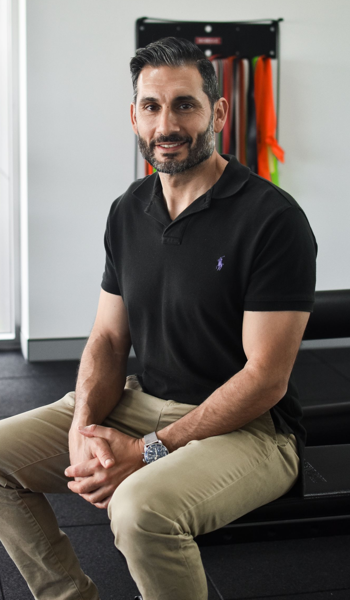 Man in black polo shirt and khaki pants sitting, smiling. Gym setting.
