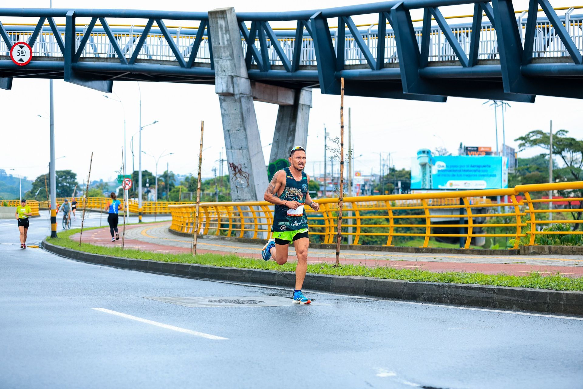 Runner in green shorts racing on a wet road under a blue pedestrian bridge.