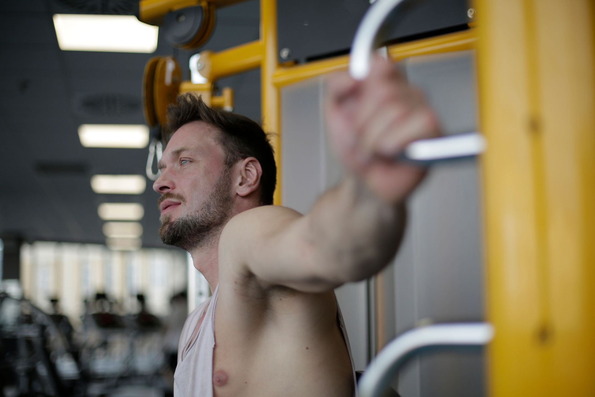 Man using a cable machine in a gym, arm extended, with yellow equipment and blurred weights in the background