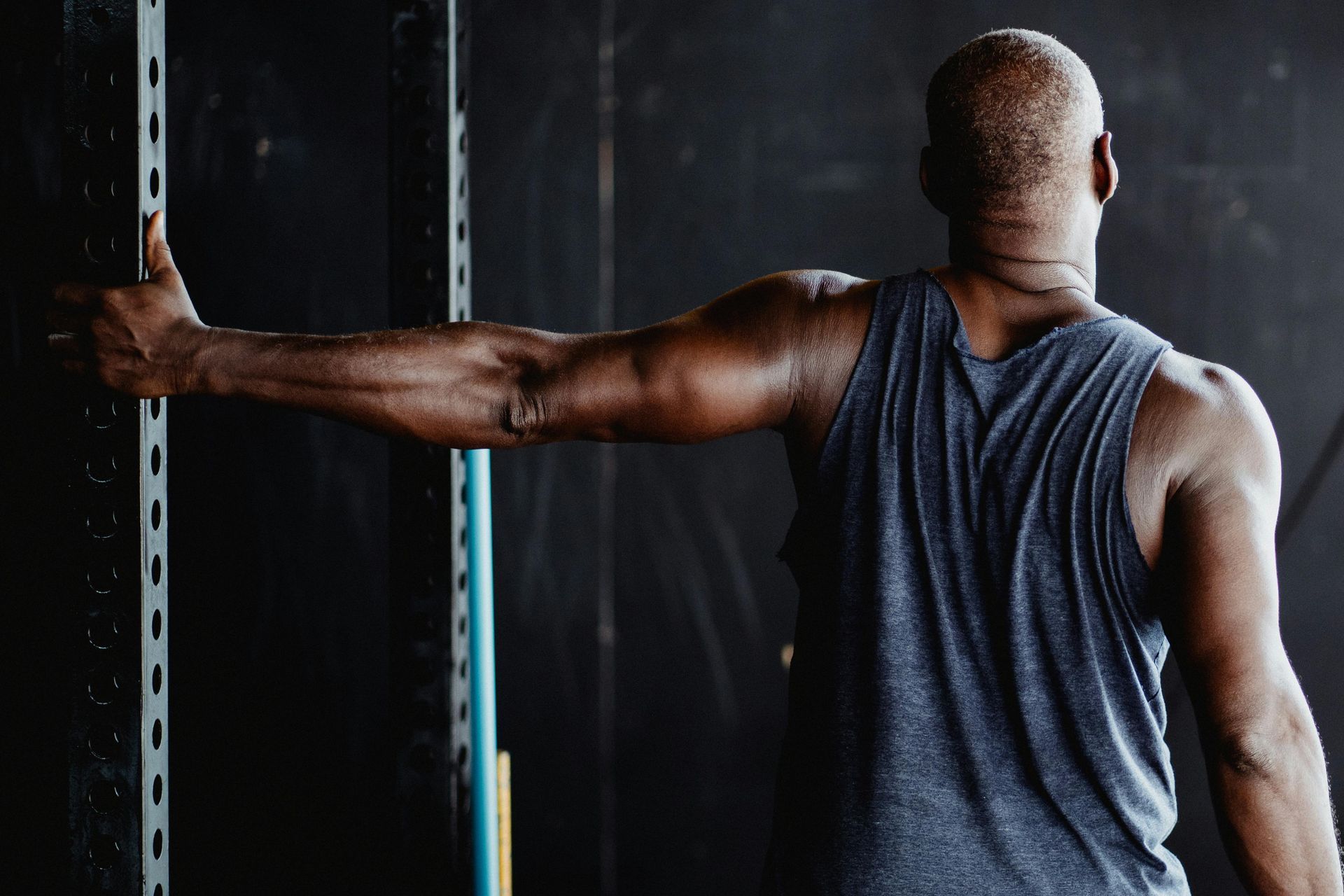 Muscular person stretching arm against a wall in a dim gym, wearing a gray tank top.