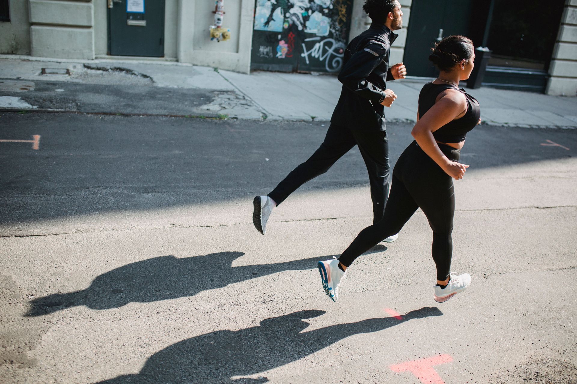Two runners in black athletic wear sprint across a sunlit street, casting long shadows.