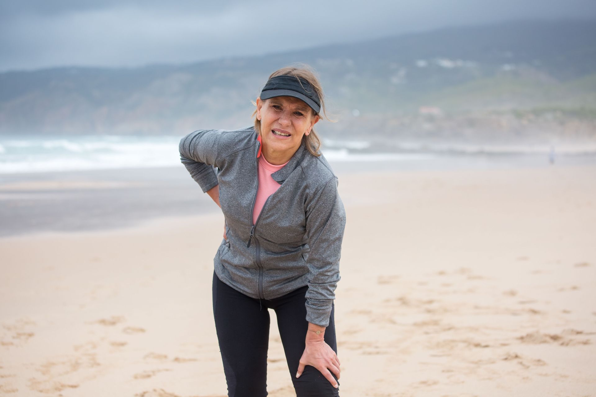 Woman in a gray jacket and cap stands on a beach, smiling with waves and cloudy hills behind her.