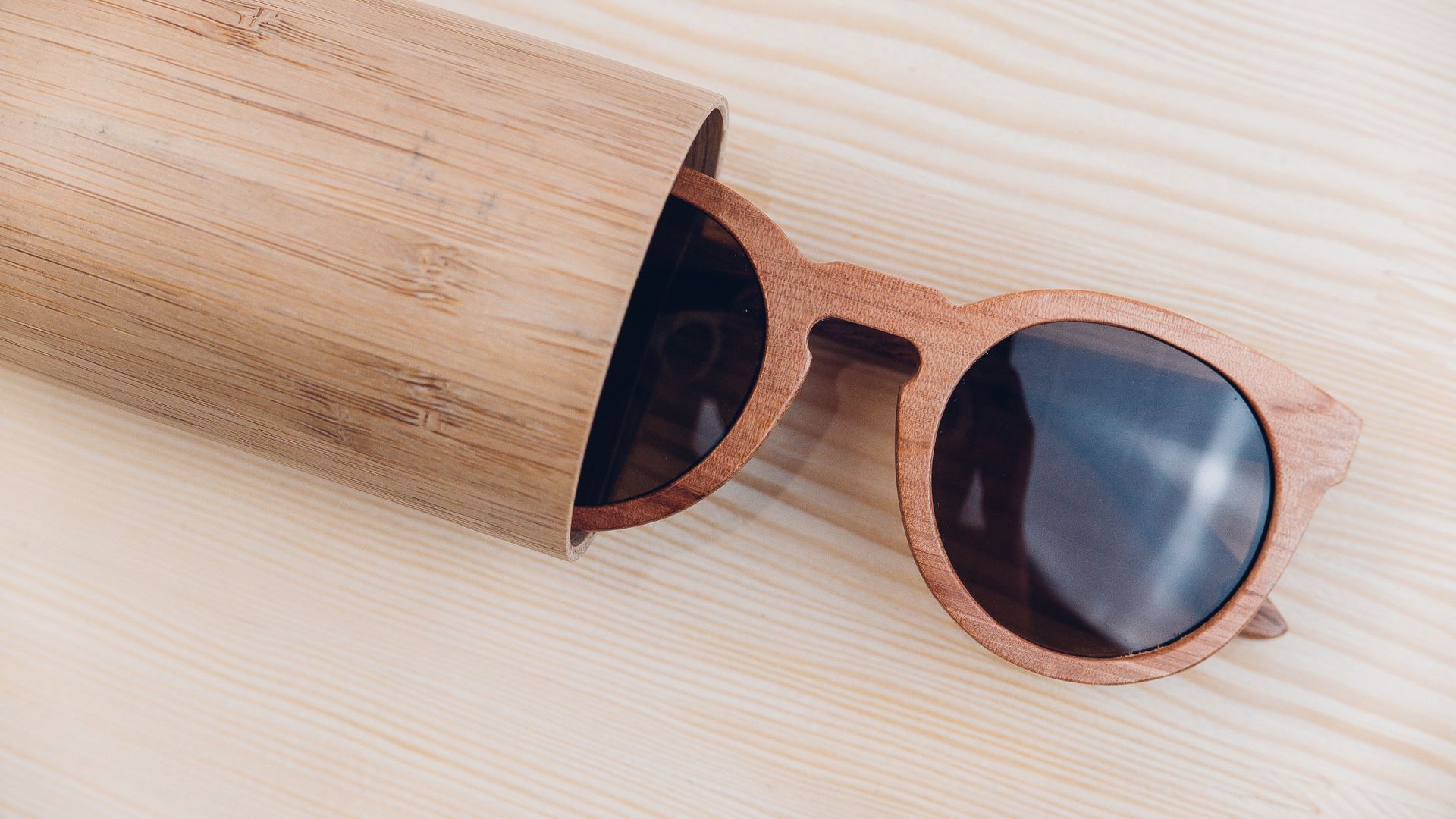 Wooden sunglasses with dark lenses beside a wooden cup on a light wood surface