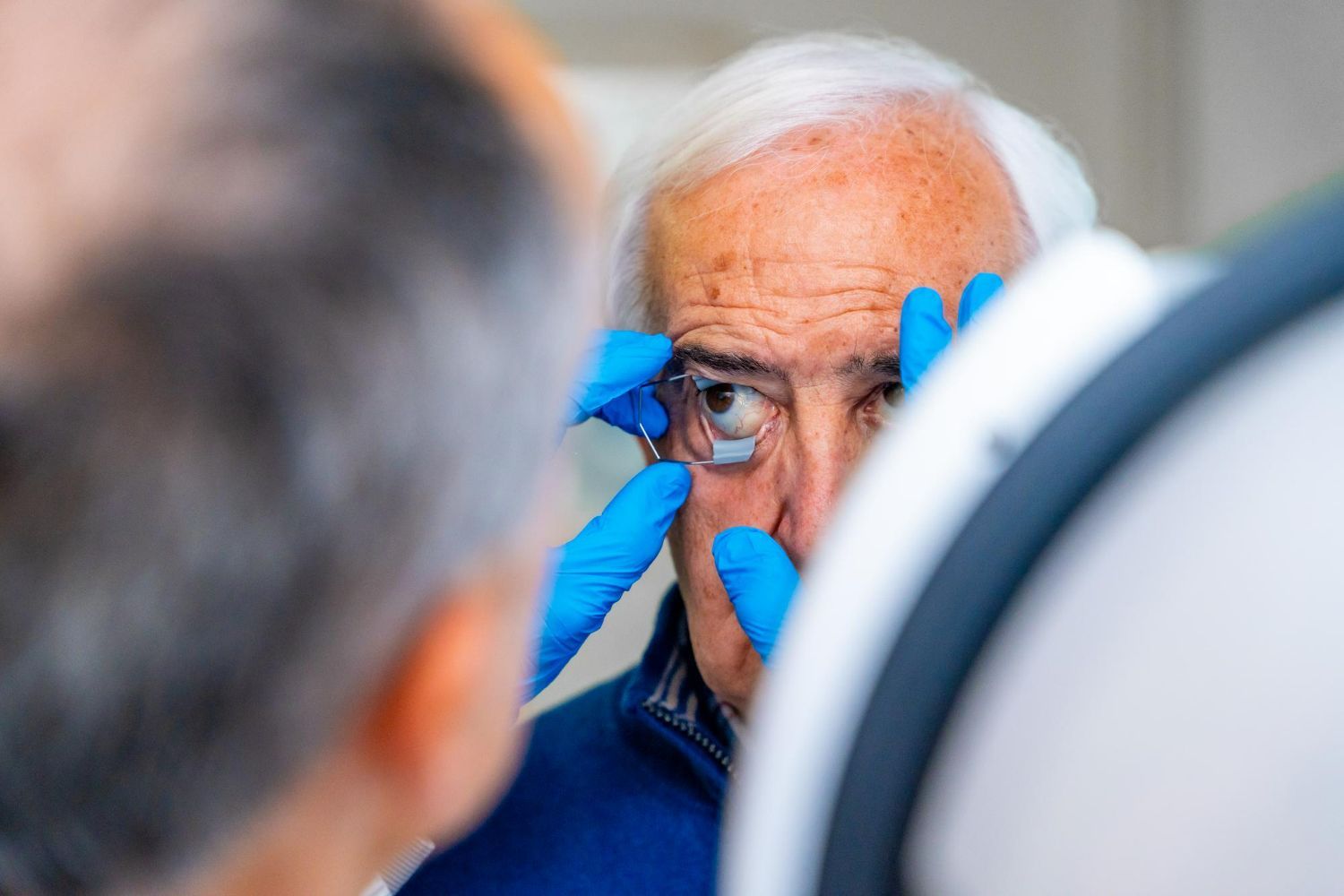 Blue-gloved hands examine an older man’s eye during a close-up medical checkup