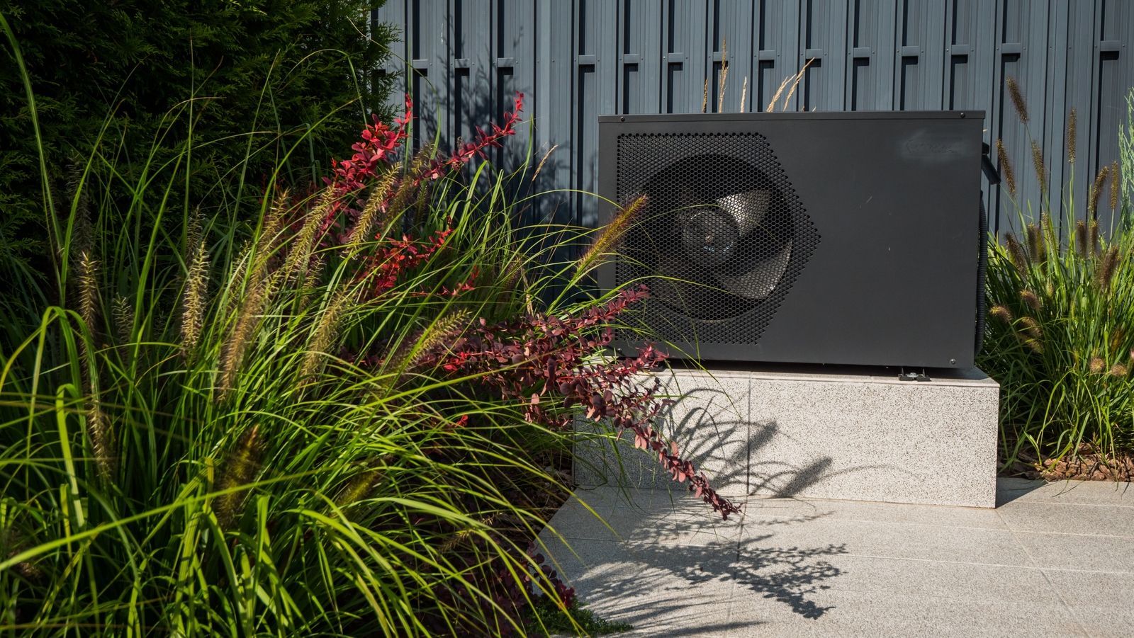 Black heat pump installed on light slabs outside, with green plants in the surroundings.