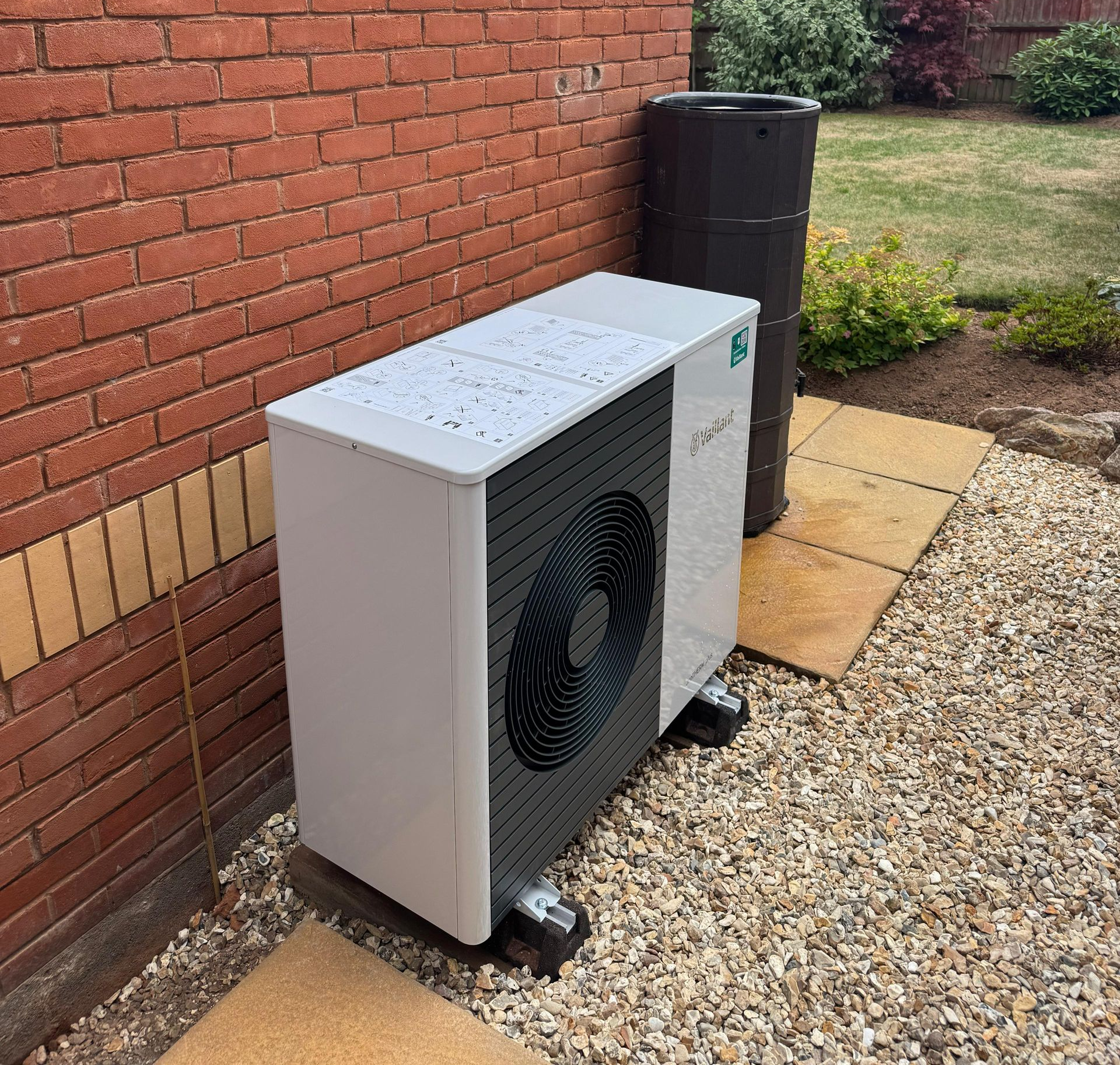 Outdoor heat pump unit beside a brick wall and black water tank on a gravel patio