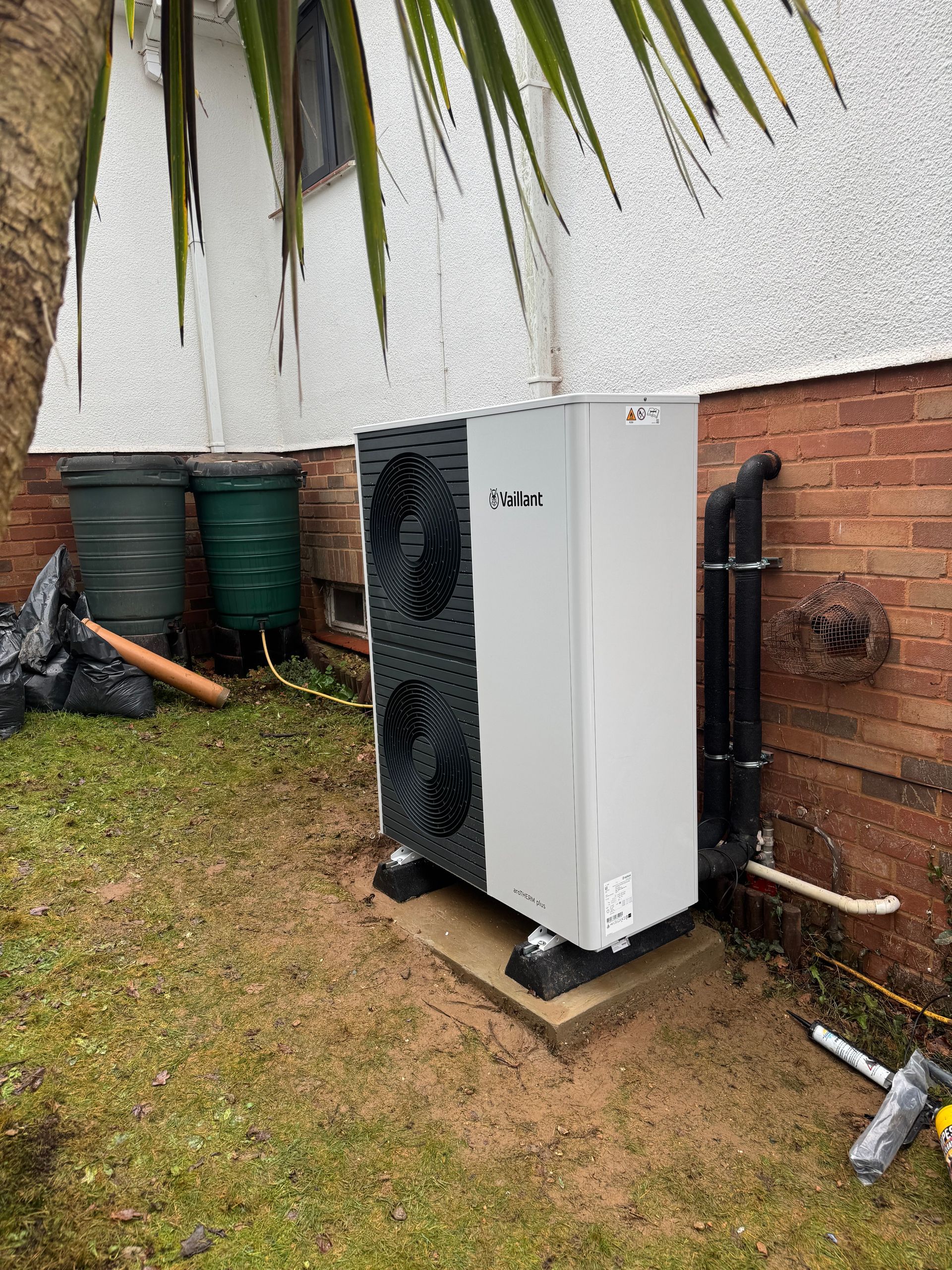 Outdoor HVAC unit beside a brick wall on a grassy yard, with pipes and utility barrels nearby