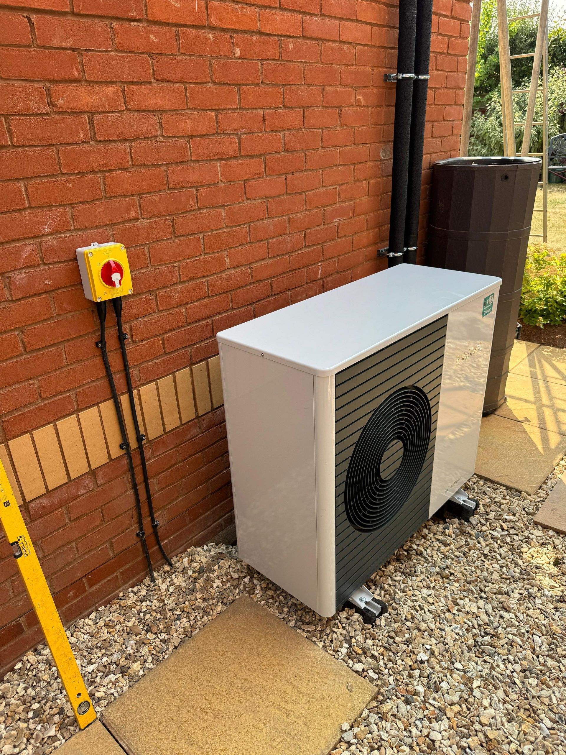 Outdoor heat pump unit beside a brick wall, with yellow emergency switch and gravel path.