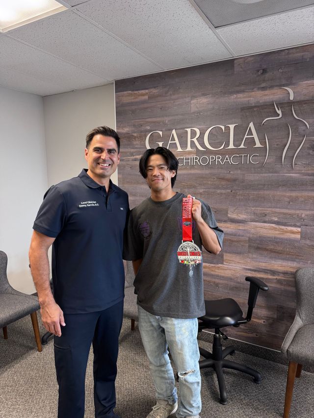 Two men standing in an office, one holding a red bottle in front of a Garcia sign.