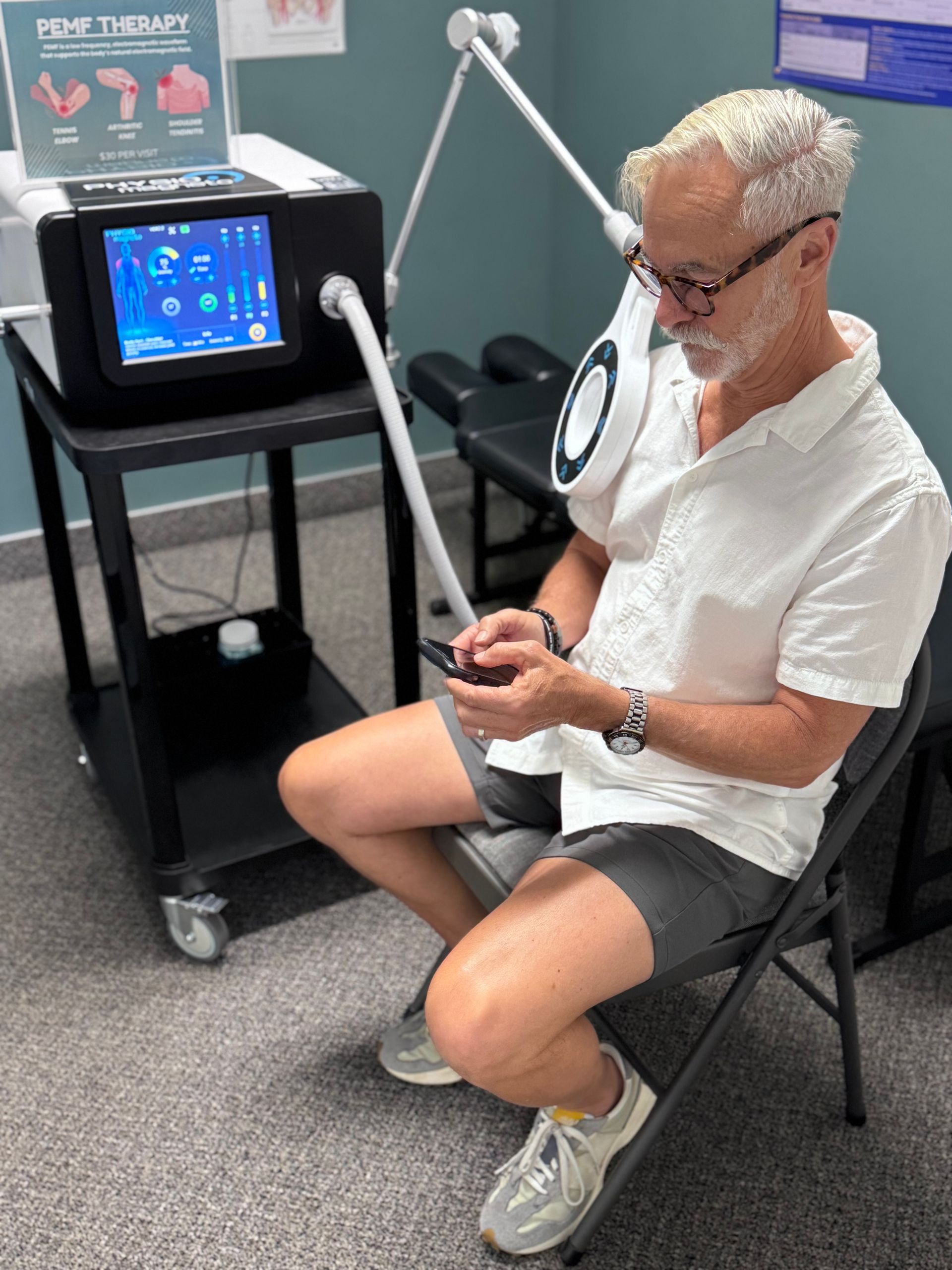 Person seated beside a PEMF therapy device, using a controller in a clinic setting