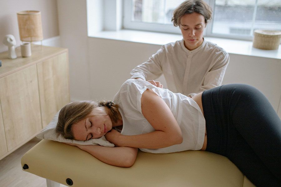 Person lying on a massage table while a therapist stands nearby in a bright room
