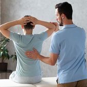 Physical therapist examining a seated patient with hands behind head, in a clinic setting