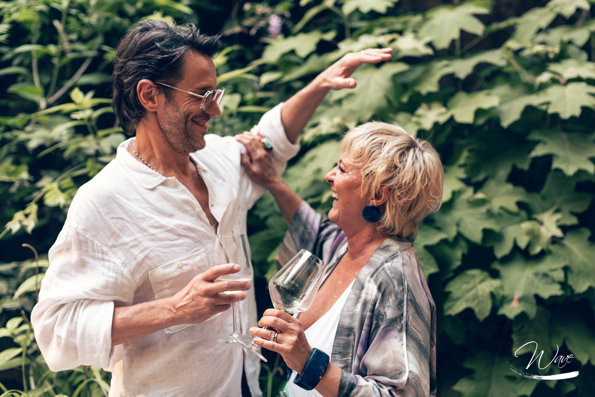 Man with arm raised, woman with wine, both laughing in lush green garden.