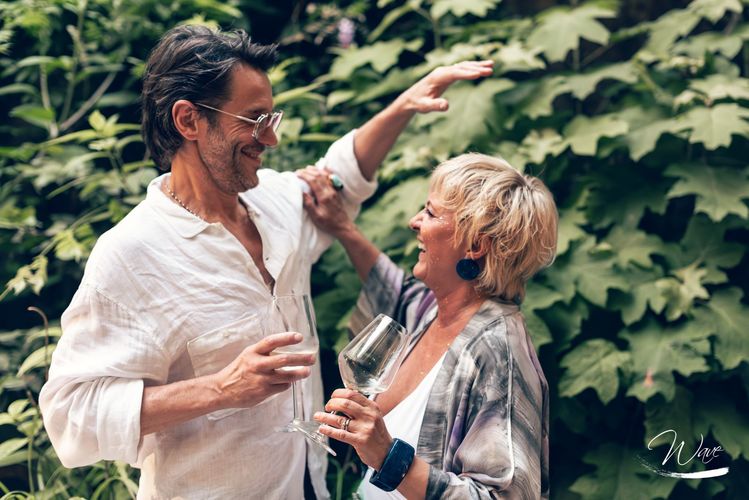 Coppia sorridente, abbracciata all'aperto, con in mano bicchieri di vino; l'uomo indica il verde.