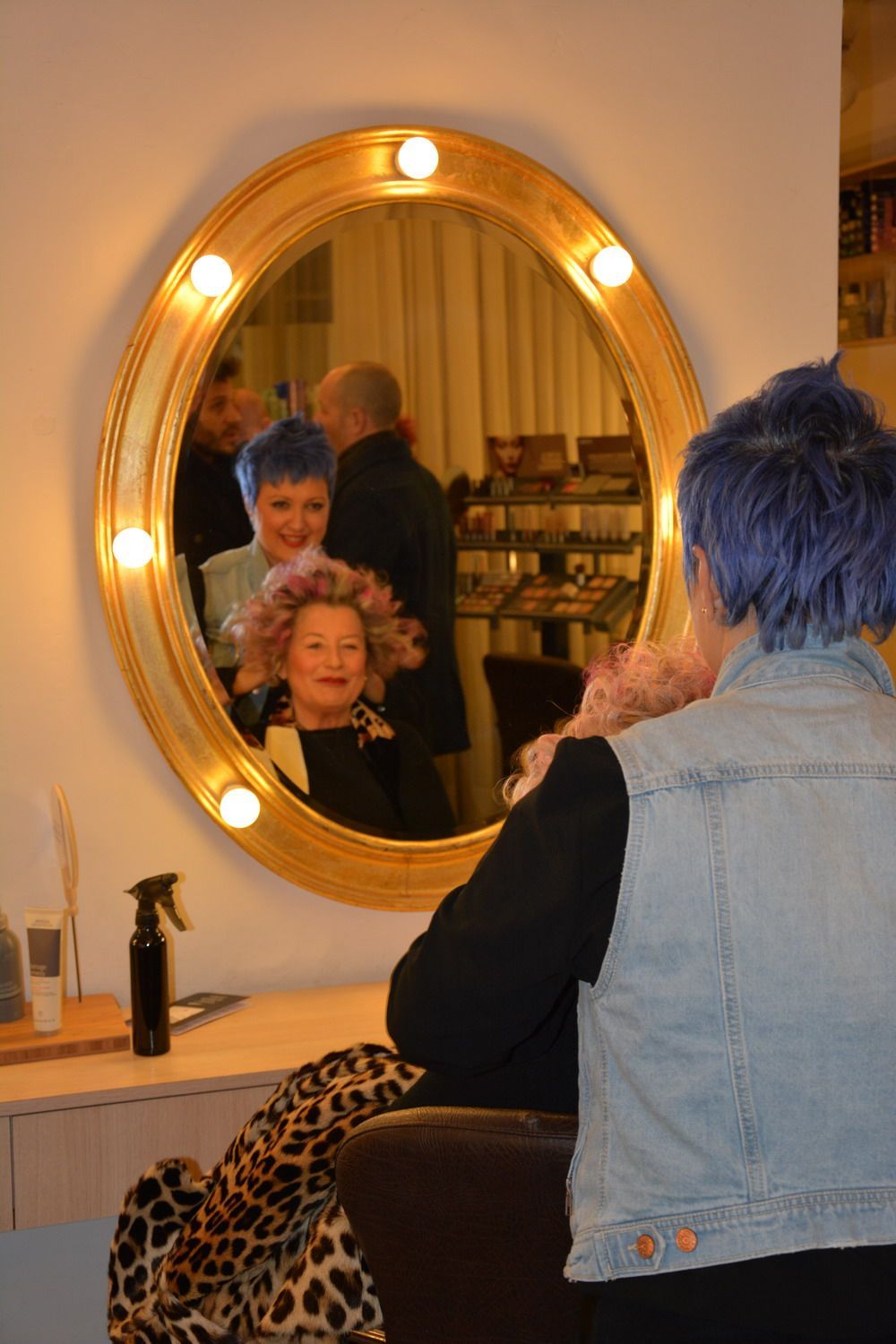 Woman with blue hair looks in a mirror at a salon, smiling at her reflection, another person also in view.