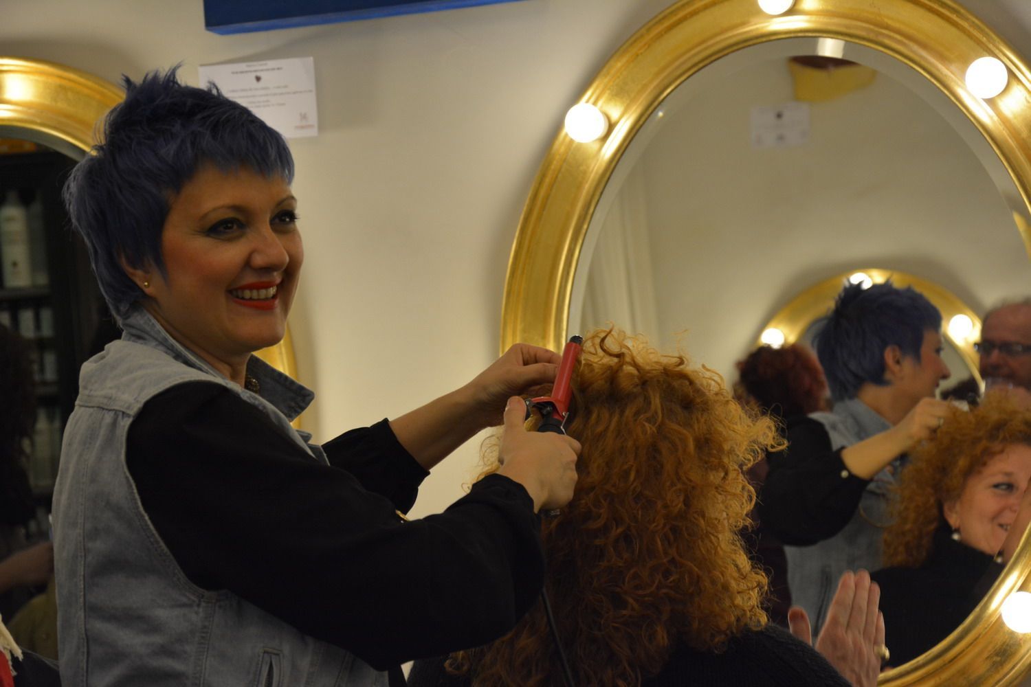 Hair stylist with blue hair curls a client's red hair in a salon, reflecting in golden mirrors.