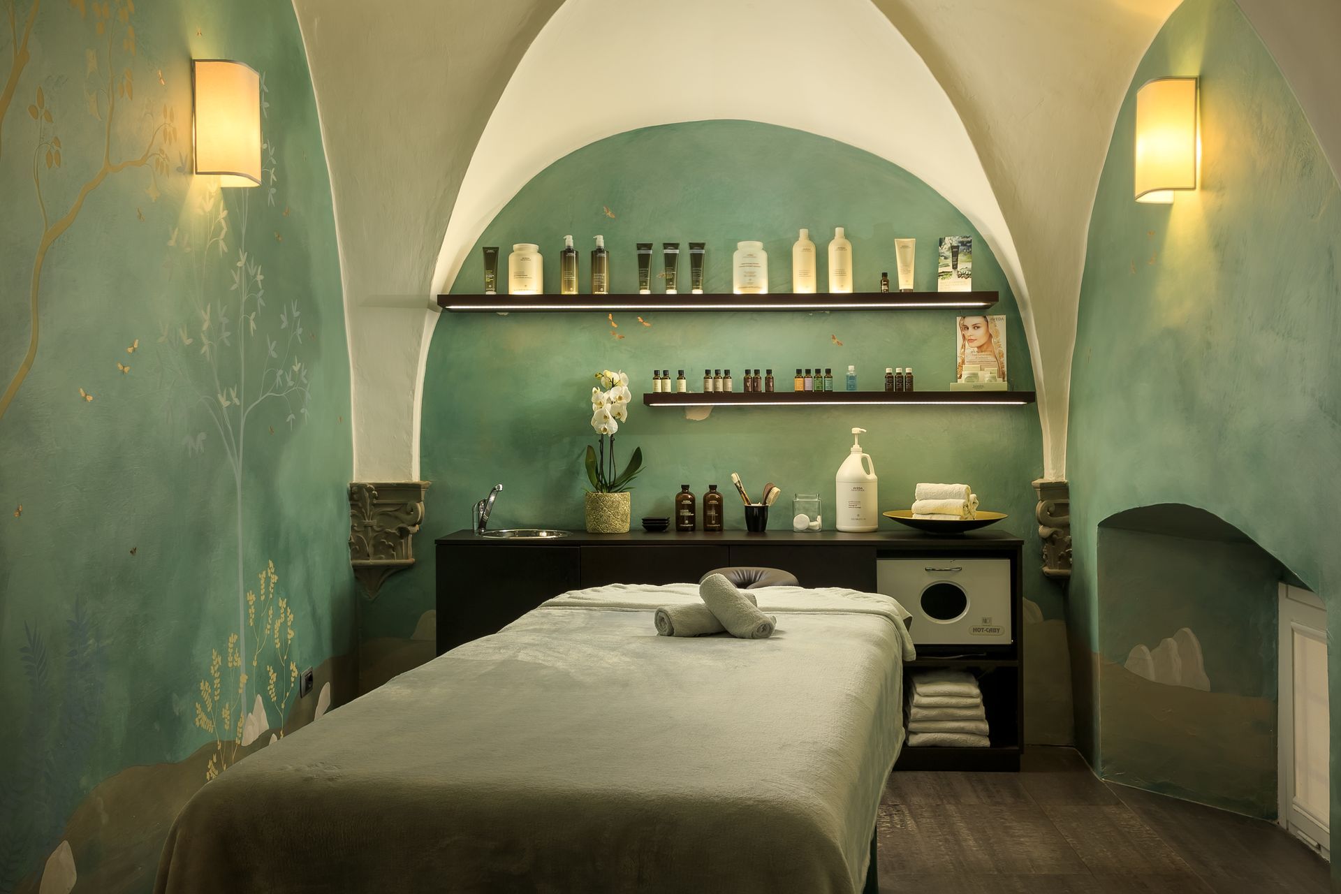 Spa treatment room with massage table, teal walls, and product shelves.