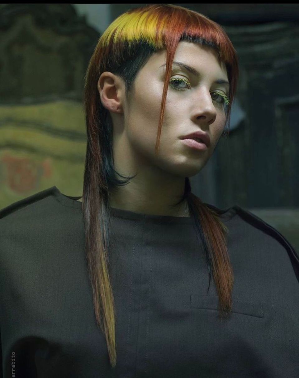 Donna con i lati rasati, una parte superiore gialla e arancione e lunghi capelli multicolori, che guarda avanti.