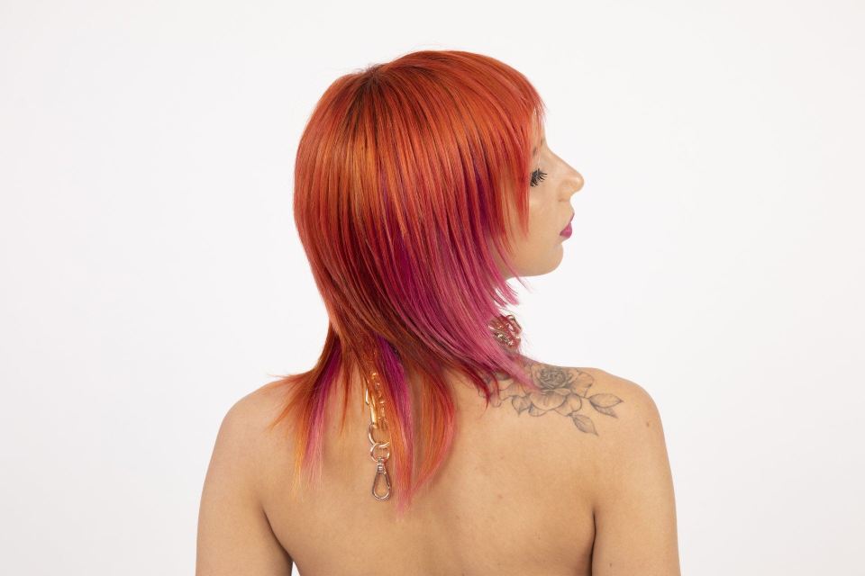 Donna con capelli rossi e rosa, di spalle, con collana e tatuaggio visibile sulla spalla.