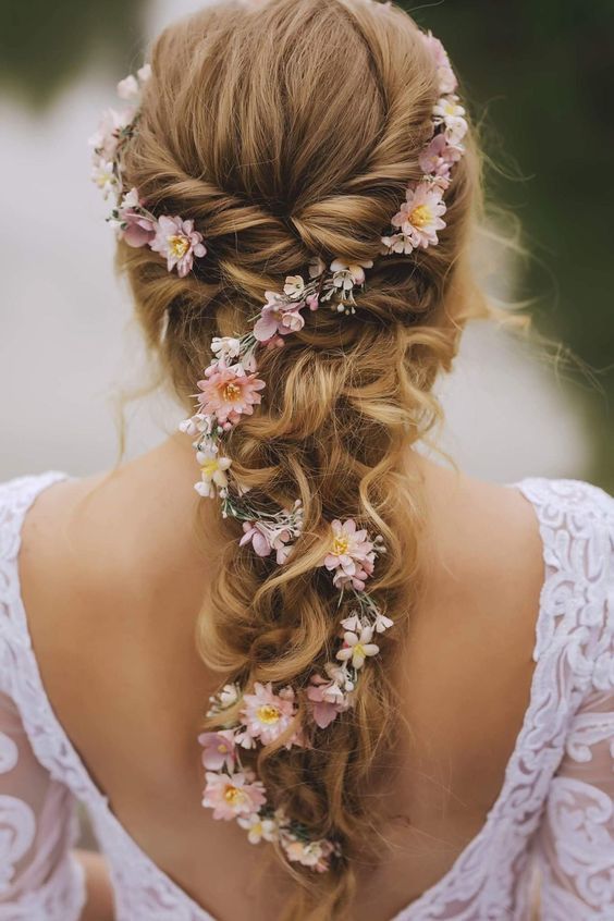Blonde woman with braided hairstyle adorned with pink and white flowers, wearing a white lace dress.