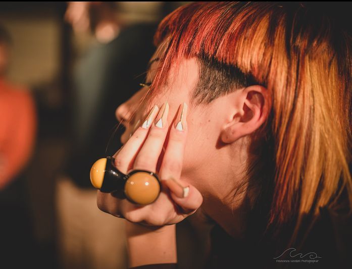 Una donna con i capelli arancioni, rasata di lato, unghie lunghe e un grosso anello si tocca il viso, in casa.