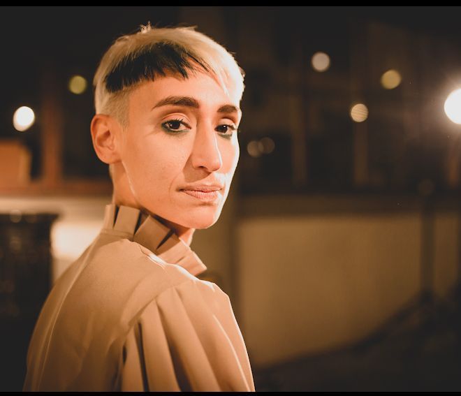 Persona con capelli biondi corti, che indossa una maglietta beige, che di notte guarda oltre la sua spalla.