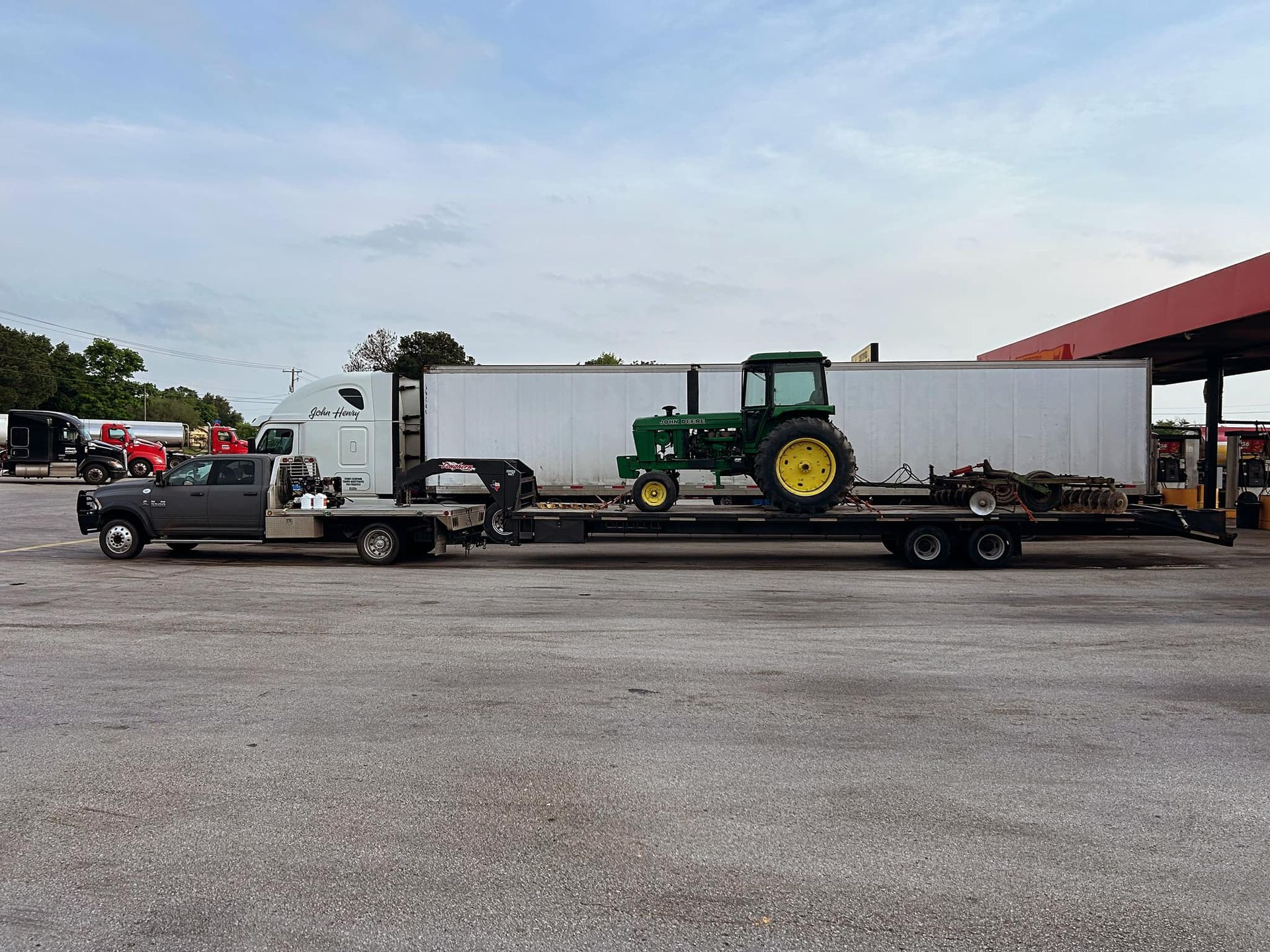 Truck hauling a John Deere tractor on a trailer at a gas station.