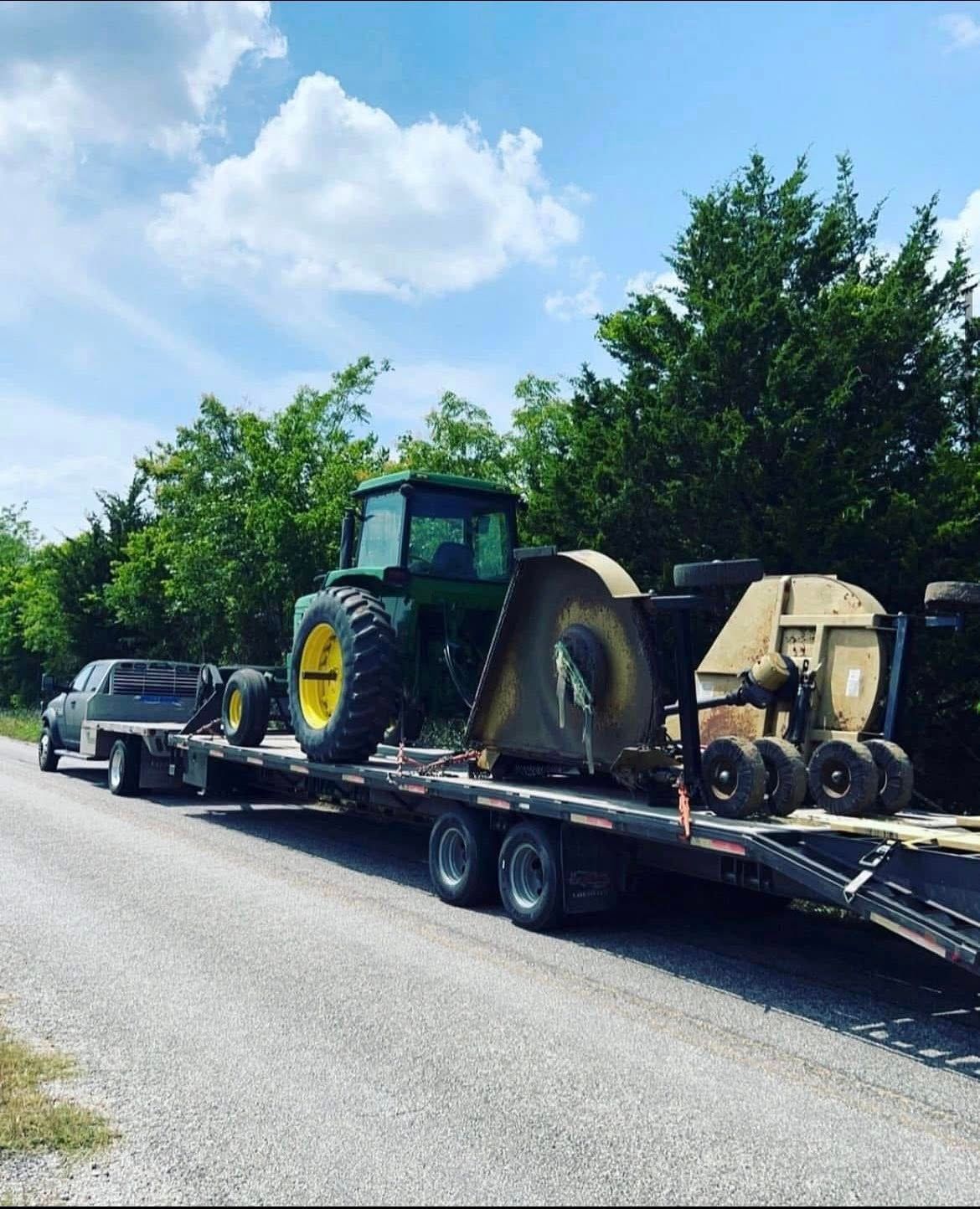 A green John Deere tractor and equipment are loaded onto a trailer, being towed down a gravel road.