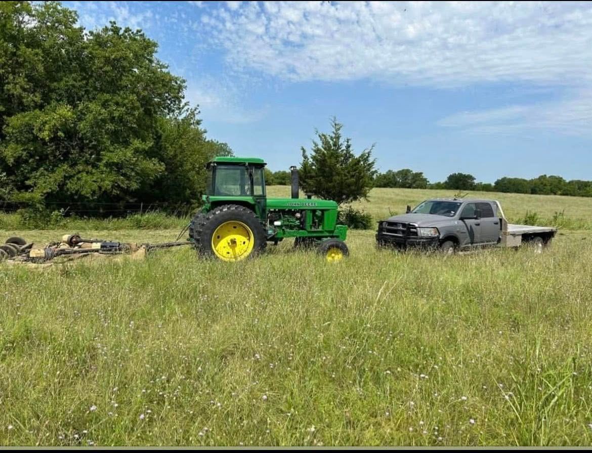 Green tractor pulling a gray truck in a grassy field on a sunny day.