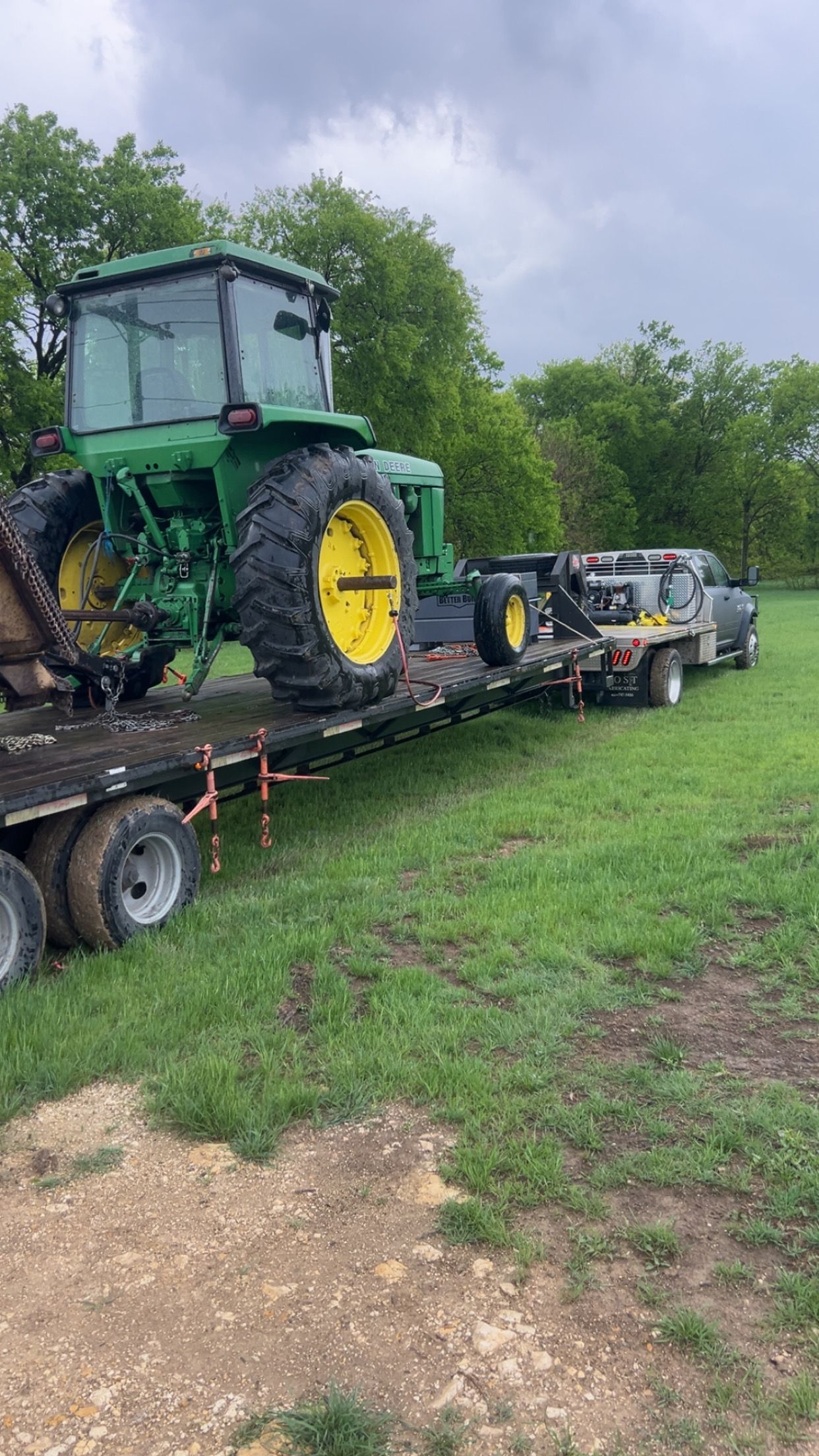 John Deere tractor on a trailer, loaded by a truck in a grassy field on an overcast day.