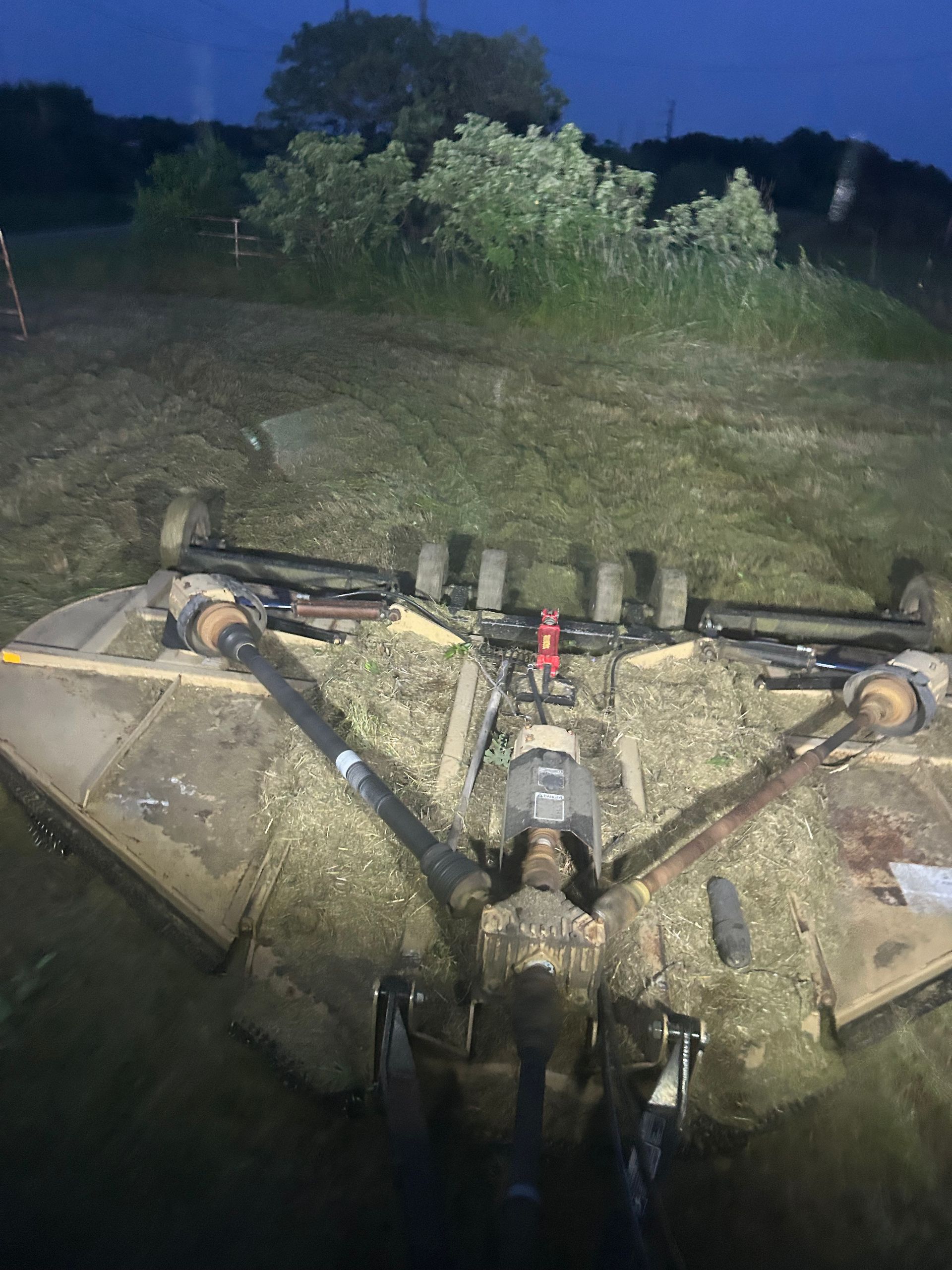A tractor-mounted mower cutting grass in a field at dusk. Blades are visible, and cut grass is strewn.