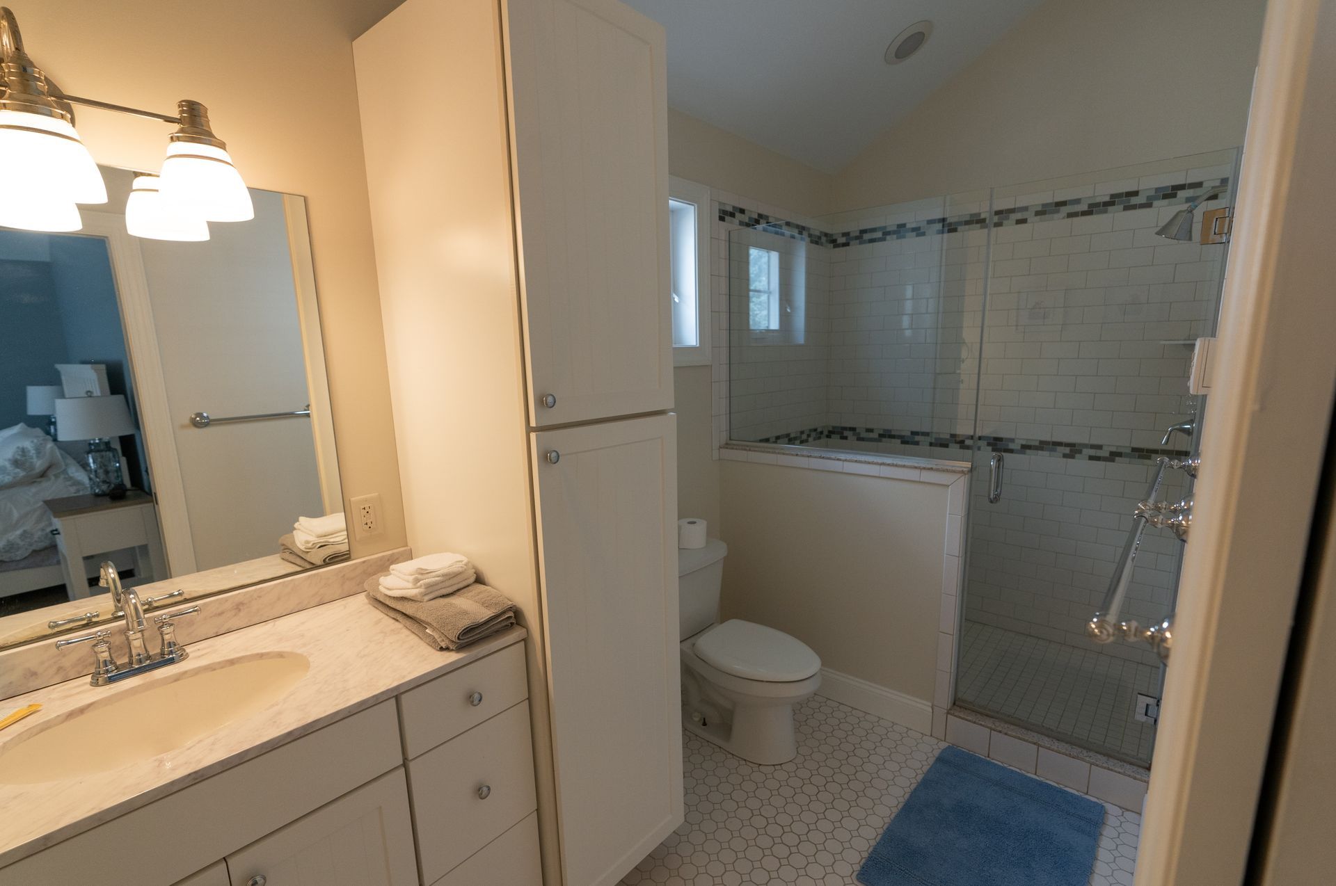 Bathroom with vanity, tall cabinet, toilet, and walk-in shower. Blue accents, beige walls, and white tile floor.
