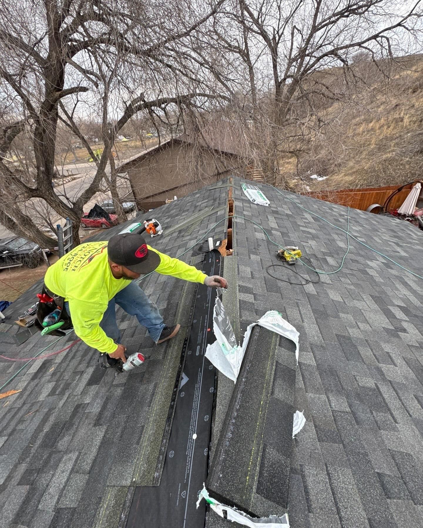 Worker in neon jacket repairs shingles on a roof with a tree-lined background