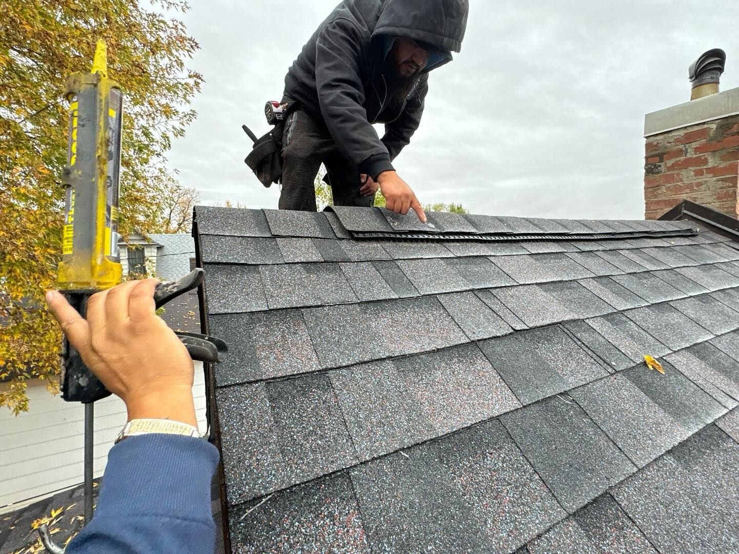 Worker installing dark gray shingles on a roof while another person measures alignment with a level.