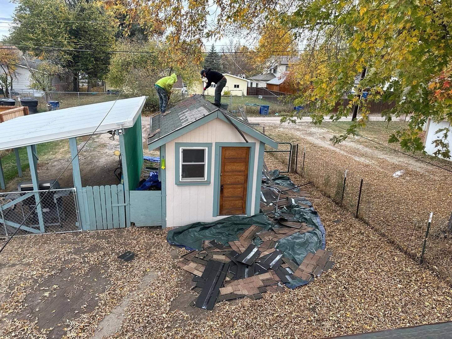 Small shed with damaged roof and scattered debris in a leaf-covered backyard, with two workers on the roof.